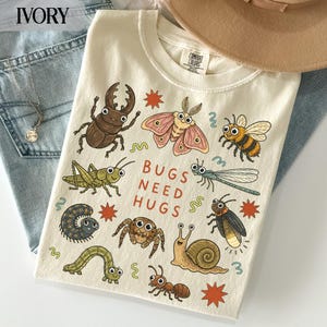 May include: An ivory t-shirt featuring a graphic print of various insects and the text "BUGS NEED HUGS" in the center. The illustration includes a beetle, moth, bee, grasshopper, dragonfly, caterpillar, spider, snail, firefly, and ant with small stars.