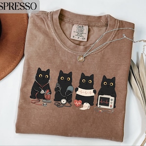 Op de afbeelding: Een espresso-kleurig t-shirt met ronde hals en een afbeelding van vier zwarte katten die medische taken uitvoeren. De katten zijn afgebeeld met een stethoscoop, bloeddrukmeter, hartritmestrook en een monitor. Voorzien van een Comfort Colors label.