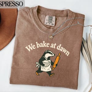 May include: An espresso-colored t-shirt featuring a graphic of a badger wearing an apron, holding a rolling pin and a bowl of dough. The text "We bake at dawn" is printed in a cream-colored font above the badger. The shirt has a crew neckline.