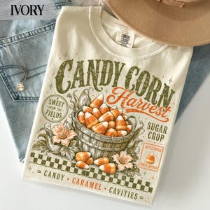 Könnte beinhalten: Ein elfenbeinfarbenes T-Shirt mit einer Grafik eines Holzkörbchens voller Zuckermais-Bonbons. Der Text lautet Candy Corn Harvest, Sweet From The Fields, Sugar Crop und Candy Caramel Cavities. Das Design enthält orangefarbene Blumen und einen Karorand.