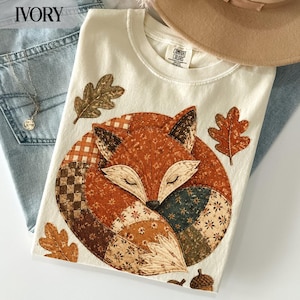 May include: An ivory t-shirt featuring a large, circular patchwork-style graphic of a curled-up fox. The fox design incorporates various brown, orange, and cream floral and checkered patterns. Three oak leaves and an acorn surround the fox.