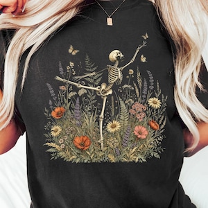 Könnte beinhalten: Ein schwarzes T-Shirt mit einem Grafikdruck eines Skeletts in Ballettpose, umgeben von einer Vielzahl bunter Wildblumen, Farnen und kleinen Schmetterlingen. Das Skelett ist in der Mitte des Blumenarrangements positioniert.
