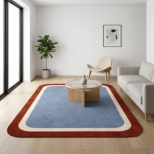 May include: A rectangular area rug featuring a large central blue panel, a thick cream-colored border, and an outer rust-red frame with rounded corners. The rug is positioned on a light-colored wooden floor in a room with white walls.