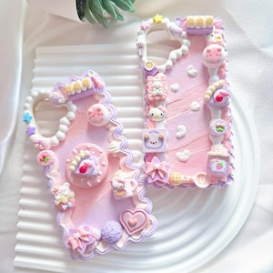 Puede incluir: Dos fundas rectangulares para teléfono con una superficie texturizada que imita crema en tonos rosa y lavanda. Los bordes están decorados con relieves estilo glaseado y varios dijes en miniatura como corazones, lazos, estrellas y figuras.