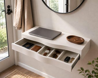 May include: A white floating wall-mounted console table with a single wide drawer divided into four compartments. The top surface holds a closed gray laptop and a small oval wooden tray with a ring. The drawer contains a passport, wallet, watch, sunglasses, car key fob, and a metal key.