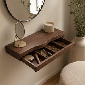 May include: A wall-mounted dark wood floating shelf with a live edge and a pull-out drawer containing four compartments. The drawer holds a makeup compact, a brush, a lipstick, and gold jewelry. A small gold-toned vanity mirror sits on top.