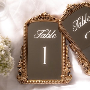 May include: Two rectangular table number signs with ornate gold-colored frames featuring intricate scrollwork and beaded edges. The center of each sign is a dark, reflective surface with the white script text Table and the number 1 printed clearly.