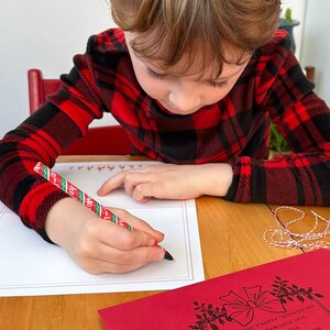 Op de afbeelding: Een kind dat een rood-zwart geruit overhemd draagt, schrijft op een witte kaart met een rood-groen gestreepte pen. De kaart heeft een rode rand en een rood-groen ontwerp langs de bovenrand.