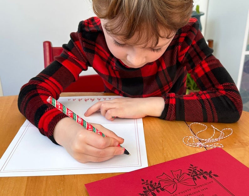 Op de afbeelding: Een kind dat een rood-zwart geruit overhemd draagt, schrijft op een witte kaart met een rood-groen gestreepte pen. De kaart heeft een rode rand en een rood-groen ontwerp langs de bovenrand.