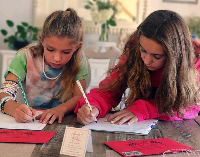 Op de afbeelding: Twee meisjes zitten aan een tafel en schrijven brieven. Ze gebruiken groene en witte potloden. De tafel heeft een houten oppervlak. Er ligt een rode envelop met een wit touwtje op de tafel. Er ligt een kaart met de tekst "Love Letters" op de tafel.