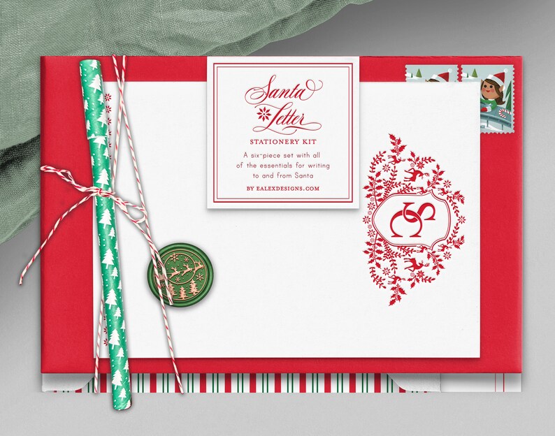Op de afbeelding: Een rode envelop met een witte kaart erin. De kaart heeft een rood-witte gestreepte rand en een rood bloemmotief met een monogram. De kaart heeft ook een wit label met de tekst "Santa Letter Stationery Kit" en "A six piece set with all of the essentials for writing to and from Santa by FALEXDESIGNS.COM".