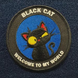 May include: A circular embroidered patch featuring a black cat with large yellow eyes and red inner ears against a blue background. The black border contains the white text "BLACK CAT" at the top and "WELCOME TO MY WORLD" at the bottom.