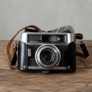 Könnte beinhalten: Eine Vintage-Kamera von Voigtländer mit schwarzer Ledertasche und braunem Lederriemen. Die Kamera hat eine silberne Deckplatte, ein zentrales Objektiv mit der Aufschrift Color-Lanthar 1:3.5/50 und Prontor 300 sowie ein strukturiertes schwarzes Gehäuse.