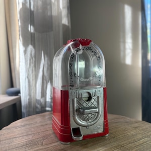 Può includere: Un distributore di gomme da masticare in plastica rossa e trasparente a forma di jukebox. La parte anteriore presenta un meccanismo a moneta in metallo argentato con una leva e un piccolo scivolo di erogazione. La parola Darmon è stampata sul vetro.