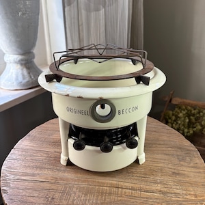 May include: A cream-coloured enamel stove with a circular metal grate on top. The front features the text ORIGINEEL BECCON with a central circular opening and three black knobs at the base. The stove has a cylindrical body with visible wear.