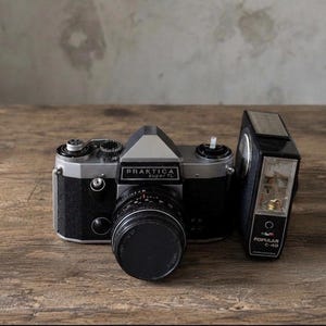Peut inclure: Un appareil photo argentique 35 mm Praktica super TL noir et argent avec un bouchon d'objectif, placé à côté d'un flash externe noir Popular C-40. L'appareil présente un viseur pentaprisme, divers cadrans et une poignée noire texturée.