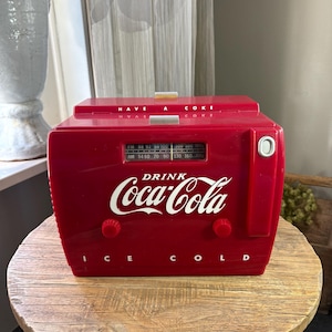 Può includere: Una radio rossa a marchio Coca-Cola a forma di ghiacciaia vintage. La parte anteriore presenta il logo bianco Coca-Cola, la scritta "DRINK" sopra e "I C E C O L D" sotto. Include un display per la sintonizzazione AM/FM, due manopole rosse e un tasto argentato.