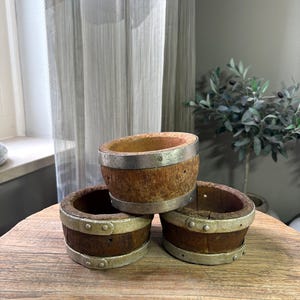 May include: Three circular wooden containers with a weathered brown finish, each reinforced by two silver-toned metal bands secured with small rivets. The containers have an open top and a hollow center, displaying a rustic texture and visible wood grain.
