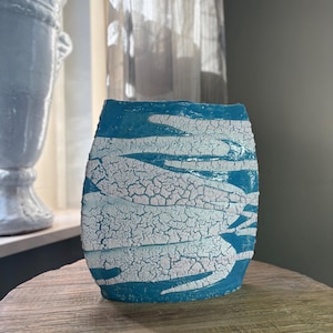 Pode incluir: Um vaso cilíndrico de cerâmica com uma cor base azul turquesa vibrante e uma camada de esmalte branco craquelado. A superfície apresenta uma rede distinta de fissuras finas sobre as áreas brancas, criando uma aparência texturizada.