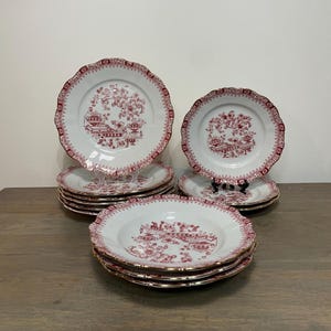 Op de afbeelding: Een set witte keramische borden en kommen met een geschulpte rand en een gedetailleerd bordeauxrood bloemenpatroon. Het ontwerp bevat ingewikkelde botanische illustraties en decoratieve randen op elk onderdeel van de serviesset.