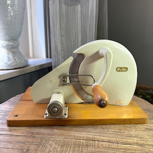 Könnte beinhalten: Ein cremefarbener, manueller Allesschneider im Vintage-Stil mit einer kreisförmigen Metallklinge und einem Holzgriff. Das Gerät ist auf einem rechteckigen Holzsockel montiert und trägt ein kleines goldenes Etikett mit der Aufschrift PeDe.