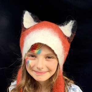 May include: A child wearing a red and white fox hat with long ear flaps. The hat is made of felted wool and has a fluffy texture. The child has rainbow face paint on their cheek.