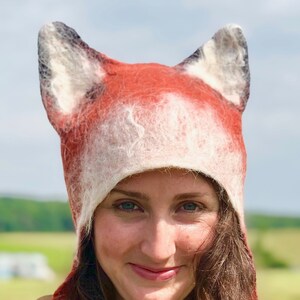 May include: A woman wearing a red and white fox hat with long brown hair. The hat has large, furry ears and a long scarf. She is wearing a white floral dress.