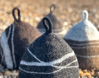 May include: Three conical felted wool sauna hats with loops at the top. The front hat is dark charcoal grey with white horizontal fibre lines. Behind it are two additional hats, one dark brown and one light beige, both featuring white line details.