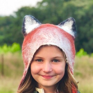 A smiling girls wears the hand felted fox hat in burnt orange with black ears and white insides and a white whisky border framing the face. Two burnt orange strips of felt act as tassels hanging down on either side of the face for tying.