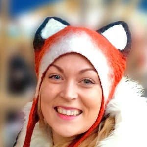 May include: A woman wearing a red and white fox hat with black ears. The hat has long ties that hang down the front of her. She is wearing a black and white polka dot shirt with a white fur collar.