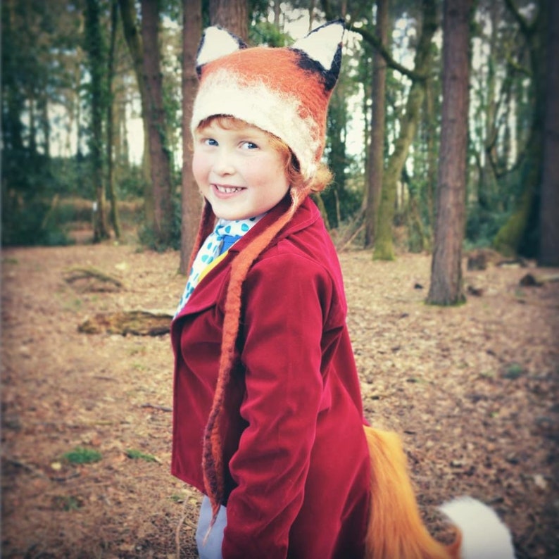 May include: A child wearing a red coat and a fox hat with a long, fluffy tail. The hat is made of felt and has a white face with black eyes and a black nose. The child is standing in a forest setting.