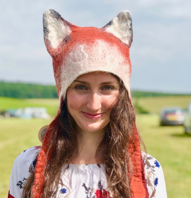 May include: A woman wearing a red and white fox hat with long brown hair. The hat has large, furry ears and a long scarf. She is wearing a white floral dress.