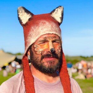 May include: A person wearing a red and white fox hat with a fox face painted on their face. The hat has long, furry ear flaps.