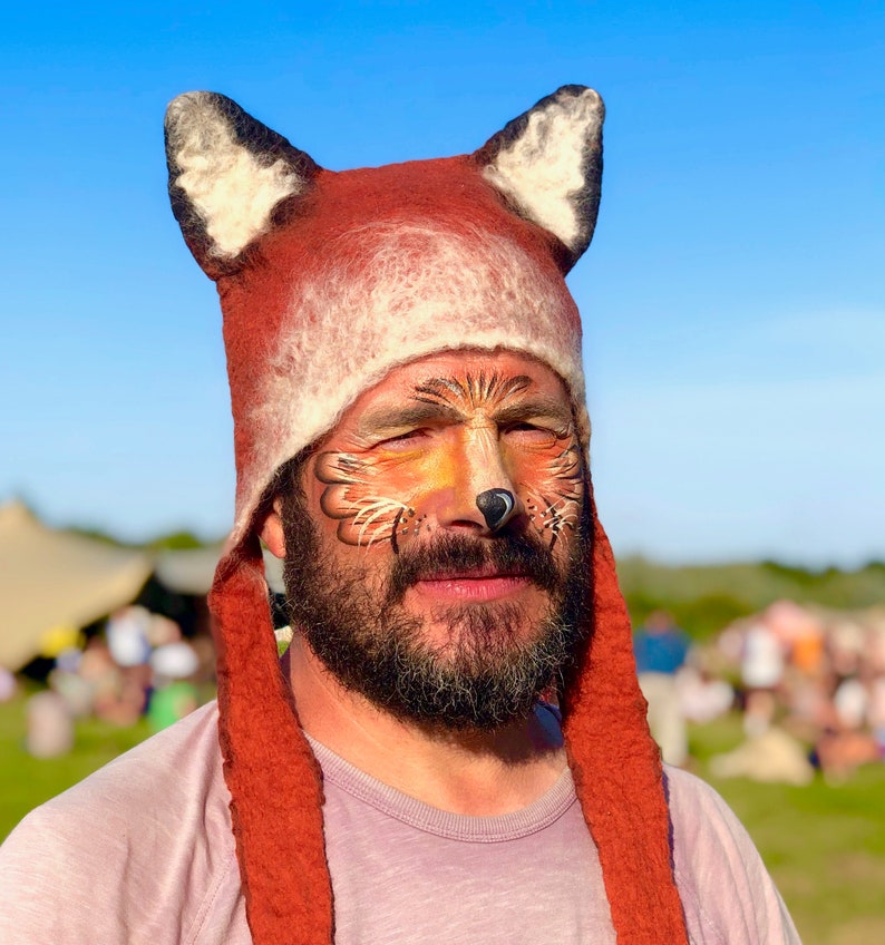 May include: A person wearing a red and white fox hat with a fox face painted on their face. The hat has long, furry ear flaps.