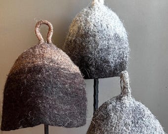 May include: Three felted wool hats in shades of grey and brown. Each hat has a rounded shape with a loop at the top. The hats are displayed on thin, black metal stands against a neutral background.