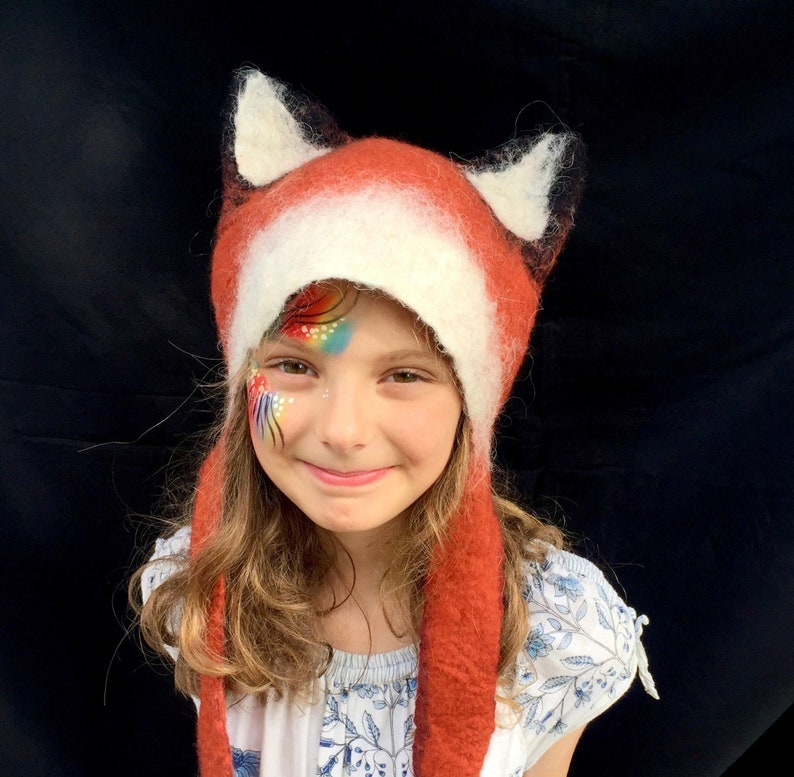 May include: A child wearing a red and white fox hat with long ear flaps. The hat is made of felted wool and has a fluffy texture. The child has rainbow face paint on their cheek.