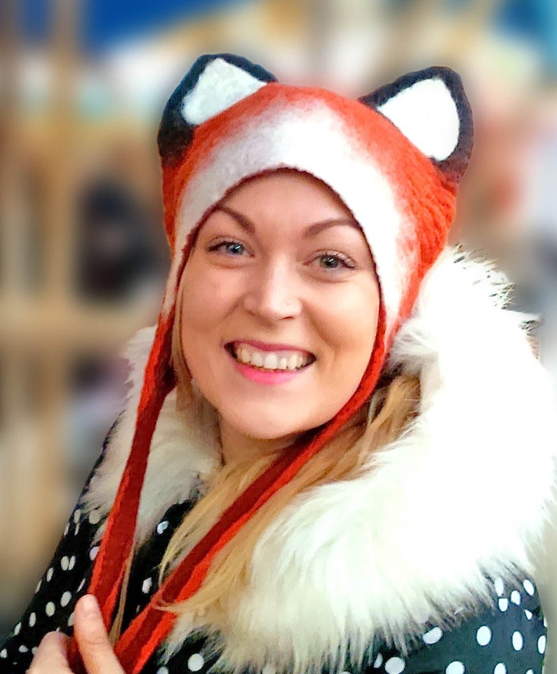 May include: A woman wearing a red and white fox hat with black ears. The hat has long ties that hang down the front of her. She is wearing a black and white polka dot shirt with a white fur collar.