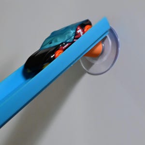 May include: A blue plastic toy car track section mounted to a wall with a clear suction cup and an orange connector. A black toy car with a teal cover and orange wheels is positioned on the track, angled downward along the blue plastic rail.