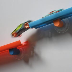 May include: A toy car in motion jumping between a blue plastic track segment and an orange plastic track segment. The tracks are mounted with small suction cups, featuring a blue support on the orange track and an orange support on the blue track.