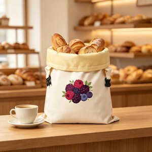 Könnte beinhalten: Ein cremefarbener Brotbeutel aus Segeltuch mit hellgelbem Innenfutter und schwarzen seitlichen Schnallen, verziert mit einer Grafik aus roten Himbeeren, dunklen Brombeeren und blauen Heidelbeeren mit grünen Blättern, gefüllt mit verschiedenen Broten.
