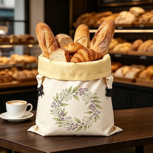 Könnte beinhalten: Ein cremefarbener Stoffbeutel für Brot mit gelbem Innenfutter und schwarzen seitlichen Schnallen, verziert mit einem botanischen Kranz aus weißen Blüten und violetten Beeren, gefüllt mit einer Auswahl an knusprigen Baguettes und goldgelben Croissants.