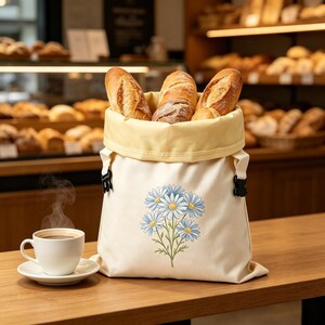 Könnte beinhalten: Ein cremefarbener Brotbeutel aus Segeltuch mit hellgelbem Innenfutter und schwarzen seitlichen Schnallen, verziert mit einer aufgedruckten Illustration von blauen und weißen Gänseblümchen. Der Beutel ist mit knusprigen Broten gefüllt und steht neben einer weißen Kaffeetasse.