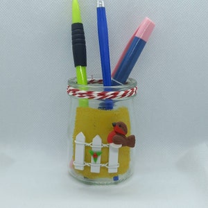 May include: A glass jar with a yellow background and a red robin bird figurine perched on a white picket fence. The jar has a red and white twine tied around the top and is filled with three pens.