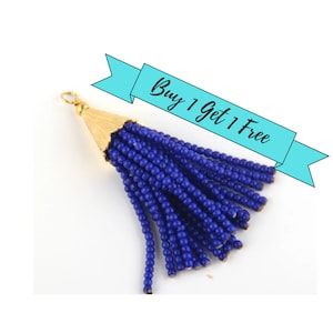 May include: A blue beaded tassel pendant featuring a gold-toned metal cap with a small circular loop at the top. The tassel consists of numerous strands of small, uniform blue beads hanging from the textured gold-coloured metal cone-shaped cap.