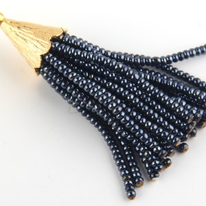 May include: A decorative tassel pendant featuring a gold-toned metal cap with a textured, fluted surface and a small circular loop at the top. The tassel consists of numerous strands of small, shiny, dark navy blue beads hanging downwards.