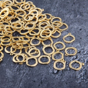 Op de afbeelding: Een grote stapel gouden metalen ringen met een getextureerd oppervlak. De ringen hebben twee kleine lussen aan de bovenkant, die kunnen worden gebruikt om sieraden of andere knutselwerkjes aan te bevestigen.