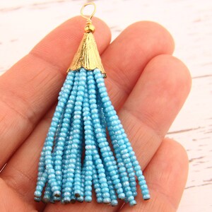 May include: A light blue beaded tassel pendant featuring a gold-toned metal cap with a textured, ribbed surface and a small circular wire loop at the top. The tassel consists of multiple strands of small, uniform, opaque light blue glass beads.