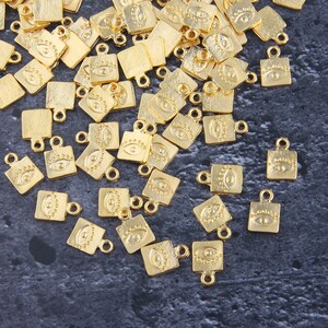 Puede incluir: Un gran grupo de abalorios de metal de color dorado en forma de cuadrado con un diseño de ojo. Los abalorios tienen un pequeño bucle en la parte superior para sujetarlos a joyas u otras manualidades.