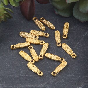 Könnte beinhalten: Eine Gruppe von 14 goldfarbenen Metall-Anhänger-Anhängern mit geometrischem Design. Die Anhänger sind in Form von kleinen, länglichen Ovalen mit einer Schlaufe oben.