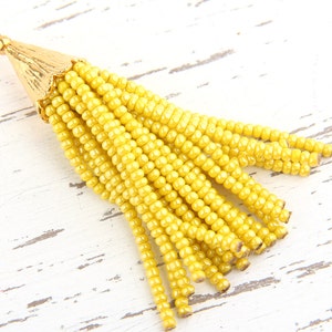 May include: A yellow beaded tassel pendant featuring a gold-toned metal cap with a textured, fluted surface and a small circular jump ring at the top. The tassel consists of multiple strands of small, uniform yellow glass beads hanging downwards.
